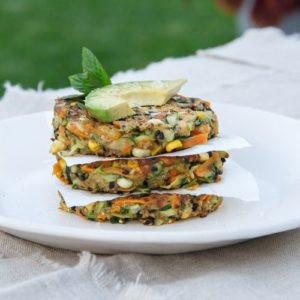 A stack of three vegan vegetable fritters on an outdoor dining table, garnished with mint and avocado