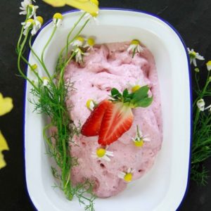 Pink ice cream in a dish with fresh chamomile flowers and sliced strawberry on top.