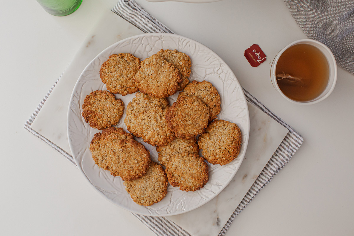 A plate full of freshly baked healthy ANZAC biscuits with a mug of hot chai tea on the kitchen counter.