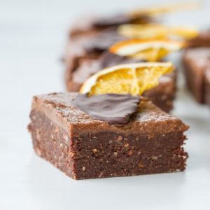 Close up of a slice of raw brownie topped with frosting and a chocolate dipped, dehydrated orange slice