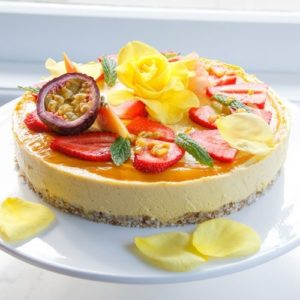 A bright yellow raw cake on a cake stand by the windowsill topped with berries, flowers and mint