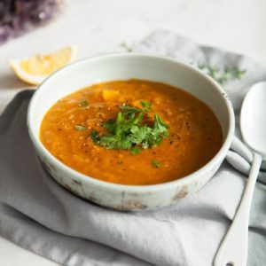 Pale green ceramic bowl full of hot red lentil soup on a blue napkin with a spoon ready to eat