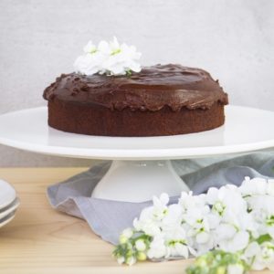 Beautiful gluten free chocolate cake covered in icing on a white cake stand surrounded by flowers