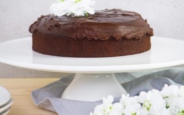 Beautiful gluten free chocolate cake covered in icing on a white cake stand surrounded by flowers