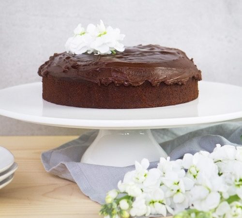 Beautiful gluten free chocolate cake covered in icing on a white cake stand surrounded by flowers