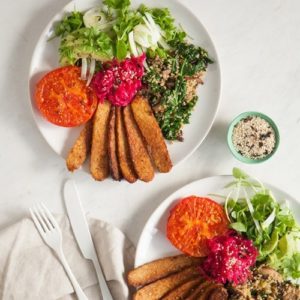 Two plates on a marble counter filled with marinated tempeh, roast tomatoes and salad greens