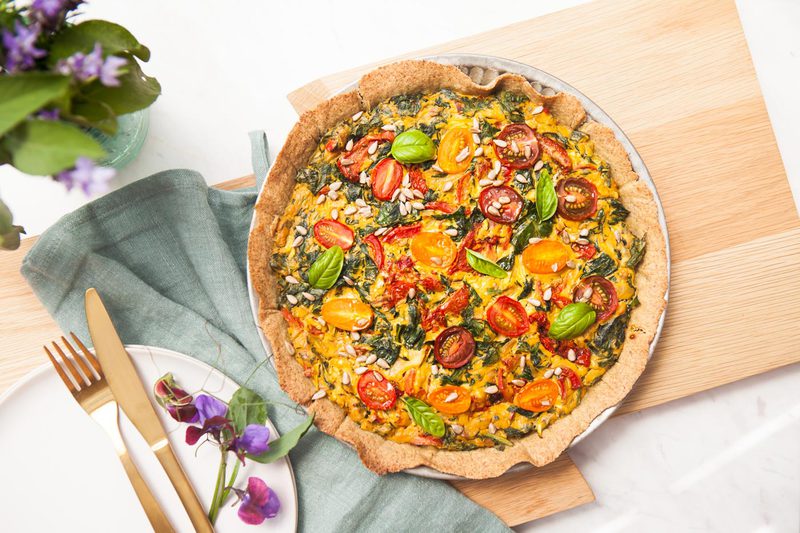 A vegan gluten free quiche on a wooden board with a rustic jar of wildflowers in an outdoor setting