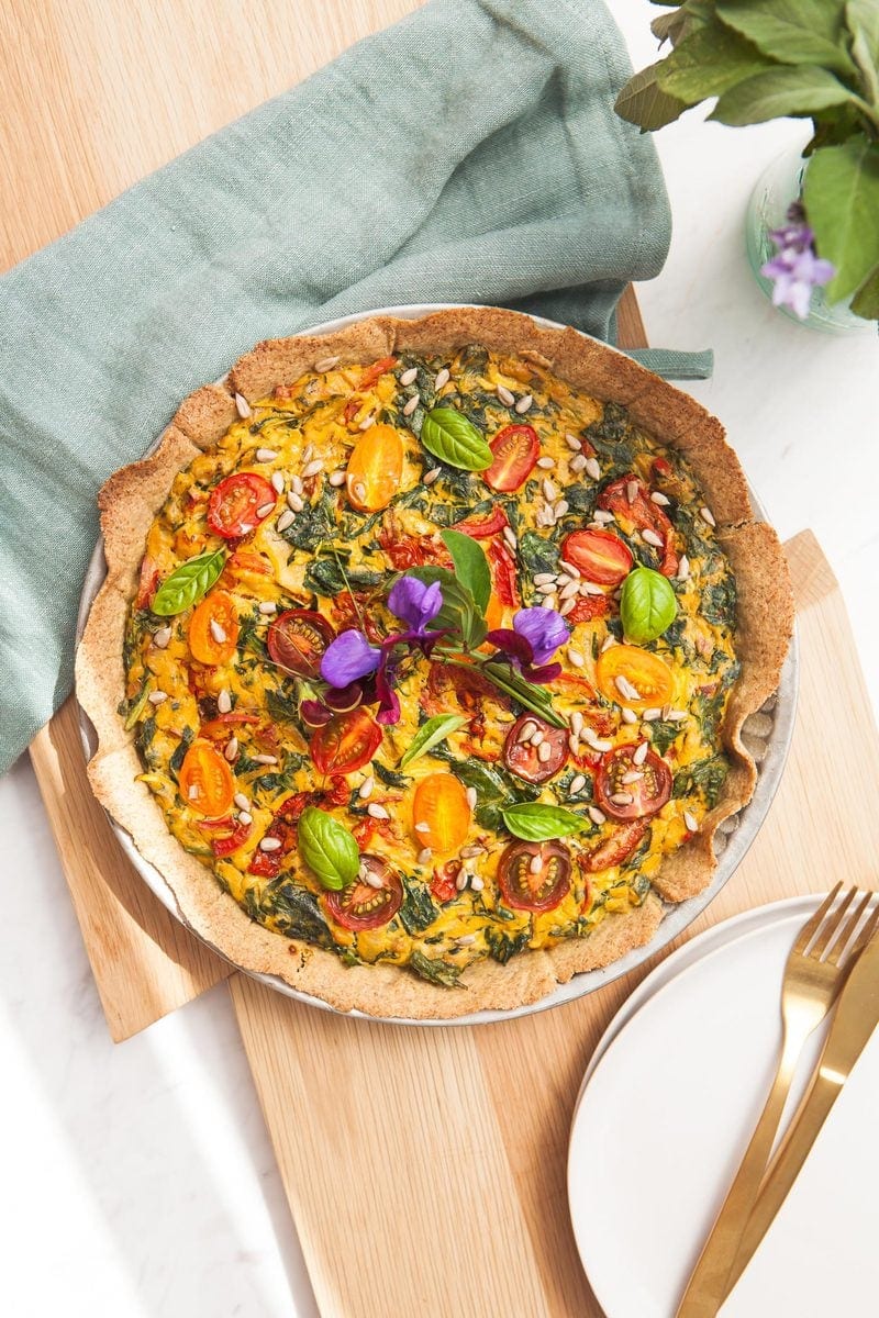 A vegan gluten free quiche on a wooden board with serving plates and gold cutlery