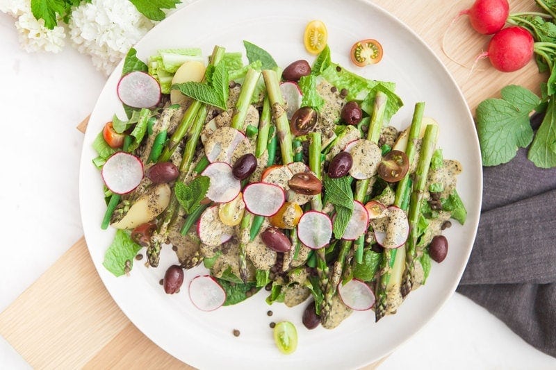 A colourful vegan Lentil and Asparagus Nicoise Salad recipe on a wooden board surrounded by fresh produce