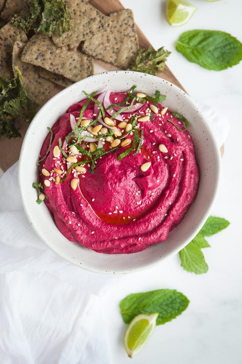 Bright pink roasted beet hummus in a bowl with a swirl of olive oil, fresh herbs on top. On a wooden board with corn chips