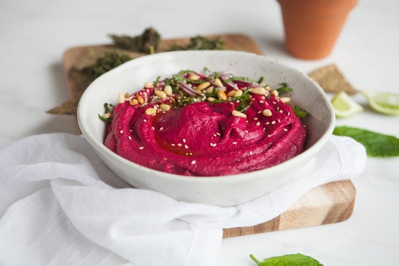 Shallow bowl filled with bright red roasted bet hummus, with pine nuts and herbs on top