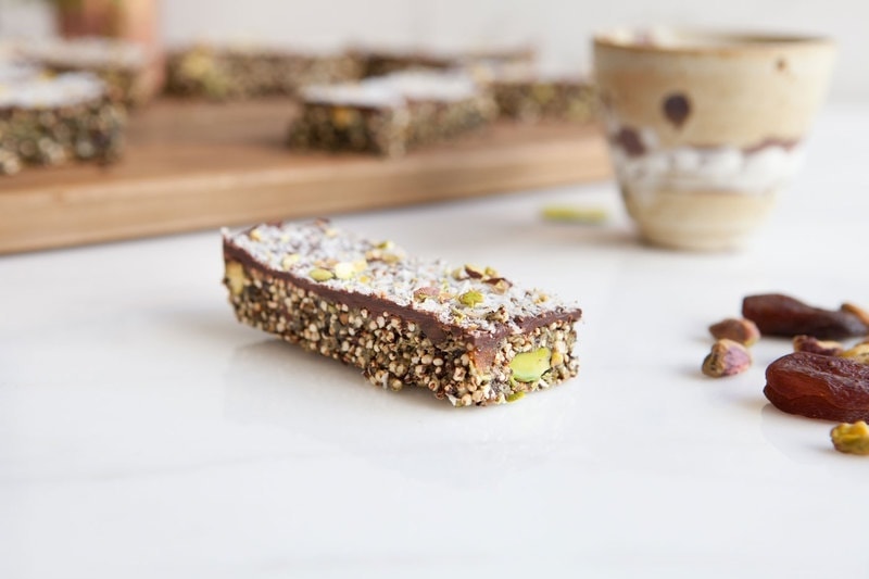 Close up of a protein packed quinoa bar with a chocolate topping, with dried apricots and pistachios beside it