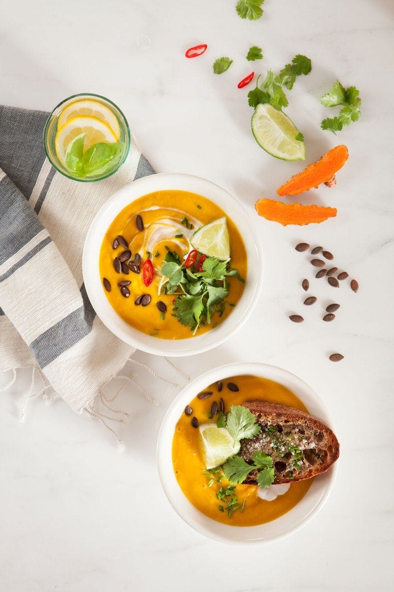 Two bowls of Anti-inflammatory Thai Pumpkin Soup surrounded by fresh ingredients - sliced turmeric, coriander leaves, sliced chilli and lime wedges