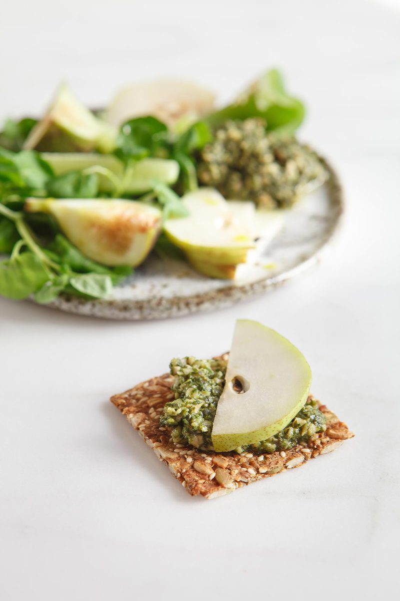 Close up of a single vegan seed cracker topped with basil pesto and a slice of fresh pear