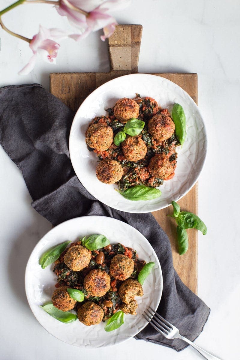 Two dinner plates filled with lentil meatballs, marinara sauce and greens, on a large wooden chopping board