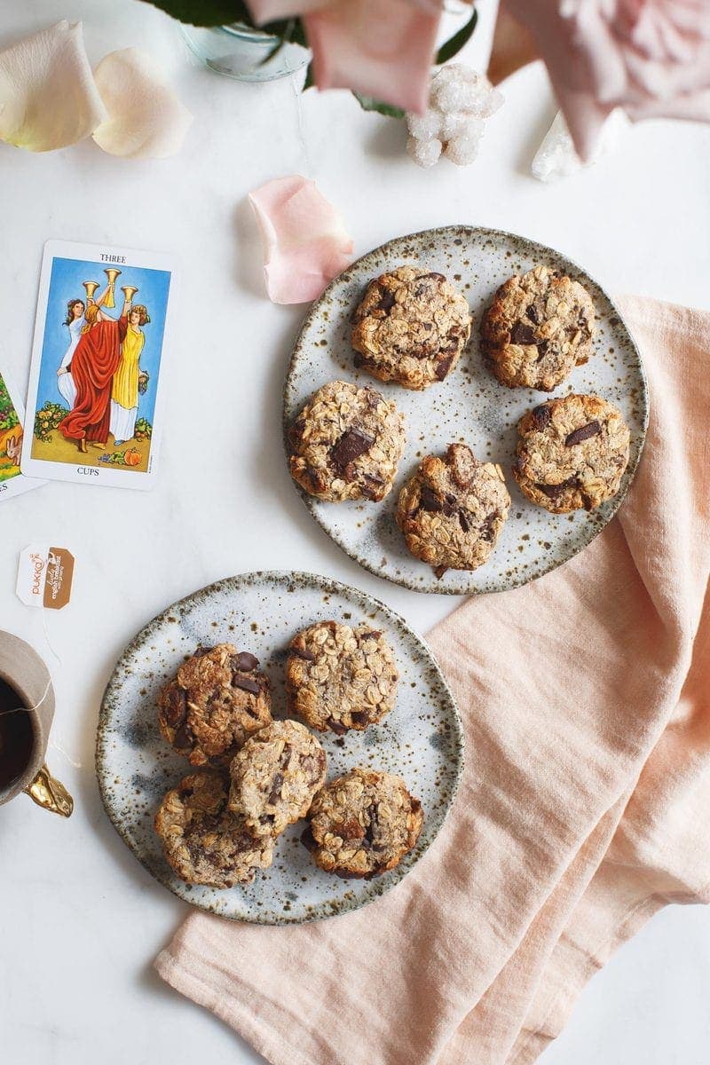 Two side plates filled with oatmeal cookies, a vase of flowers and mug of tea on marble surface