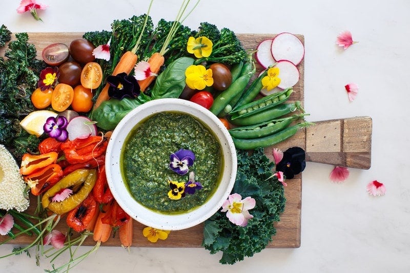 Rustic chopping board filled with coloured raw vegetables and a bowl of green, vegan basil pesto