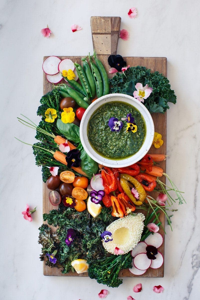 Full length rustic board on a marble counter with a rainbow of fresh vegetables and vegan hemp basil centre in a bowl