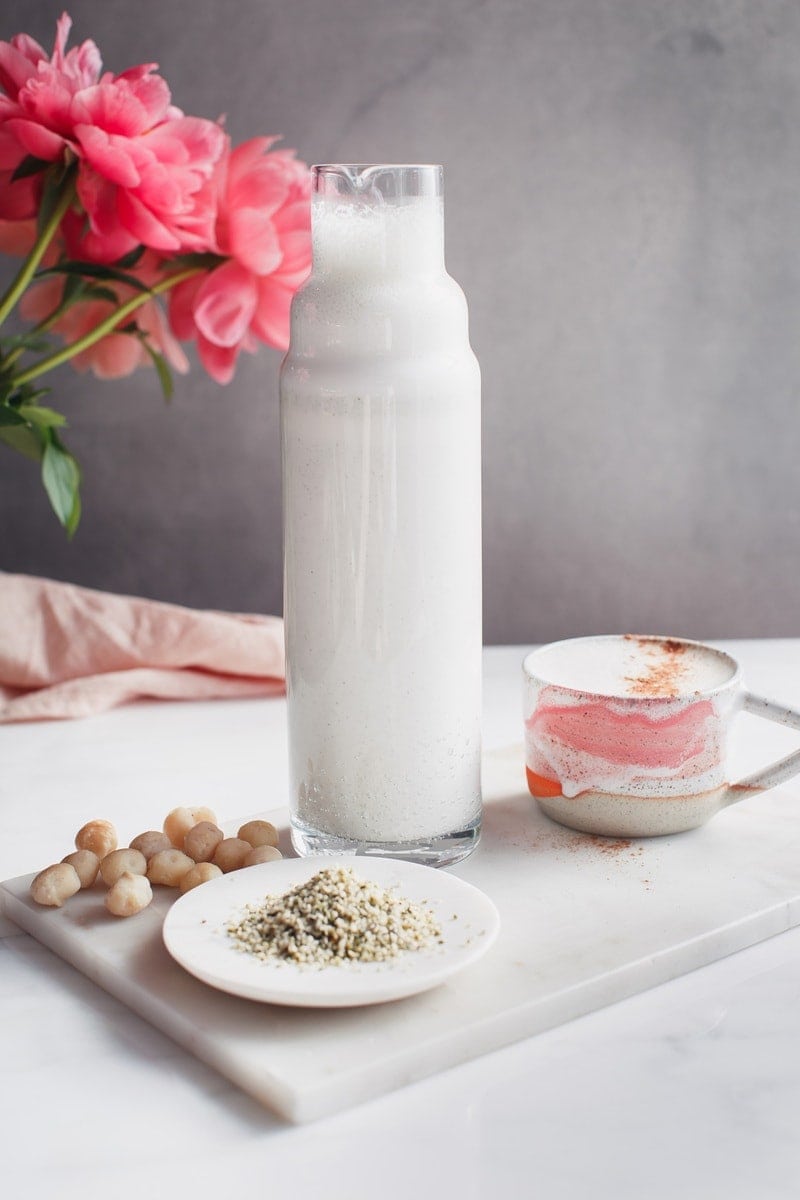 Tall modern glass jug of hemp and macadamia milk on a marble platter with vase of flowers behind