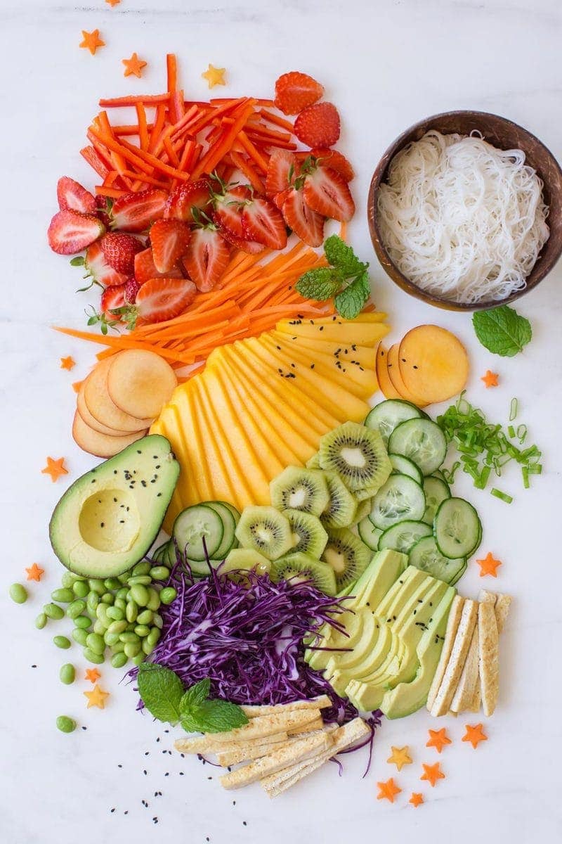 A rainbow of finely sliced fresh fruits and vegetables laid out decoratively on a marble counter, destined for summer rolls