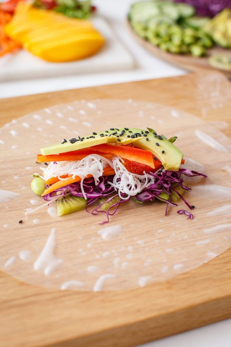 A rice paper wrapper with freshly sliced vegetables and avocado ready to be rolled, on a wooden board