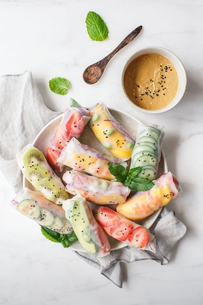 Overhead shot of 12 colourful rice paper rolls on a plate with a rich dipping sauce in a ceramic bowl next to them