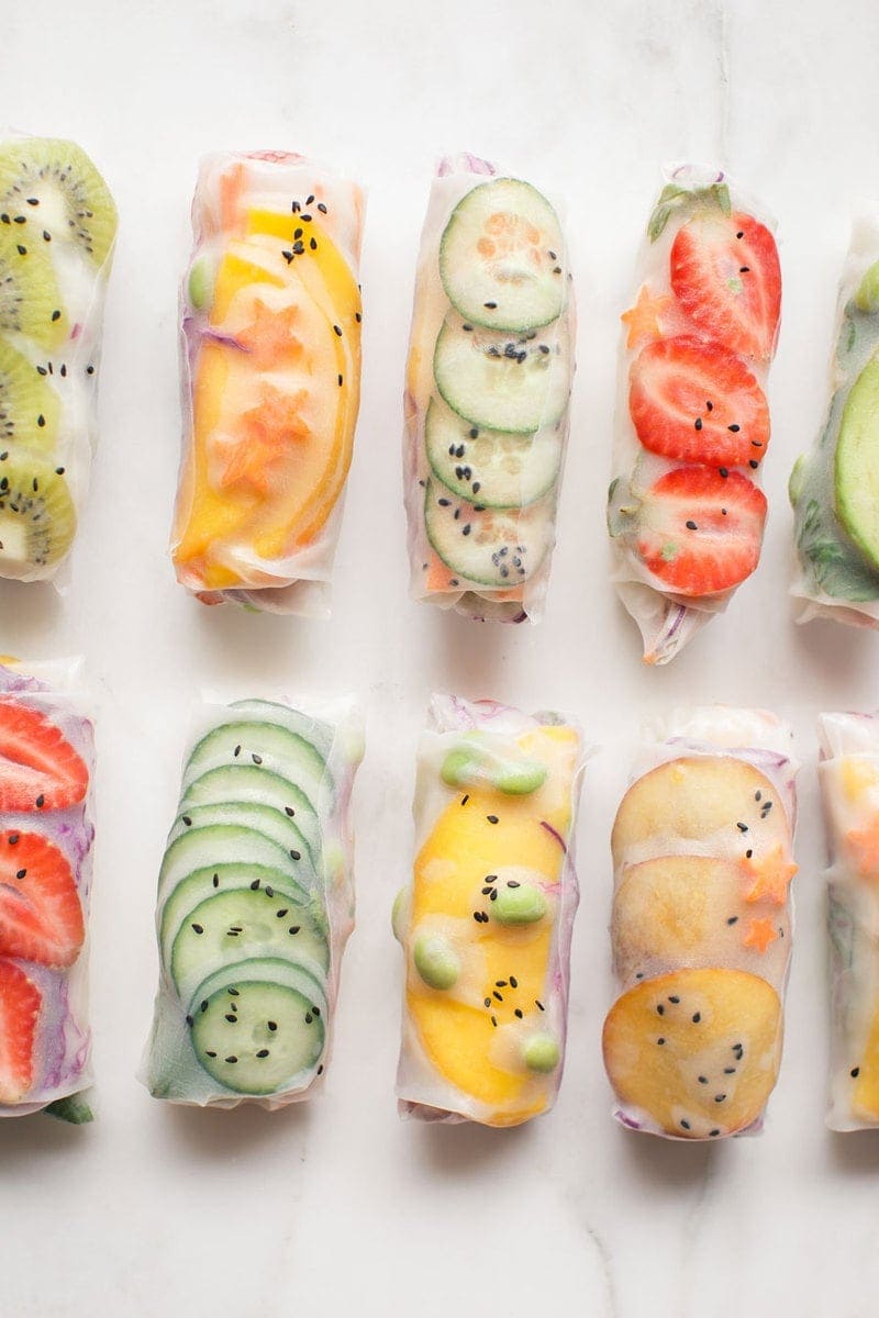 Close up of brightly coloured summer rice paper rolls filled with fruits and vegetables laid out in rows on a marble bench top