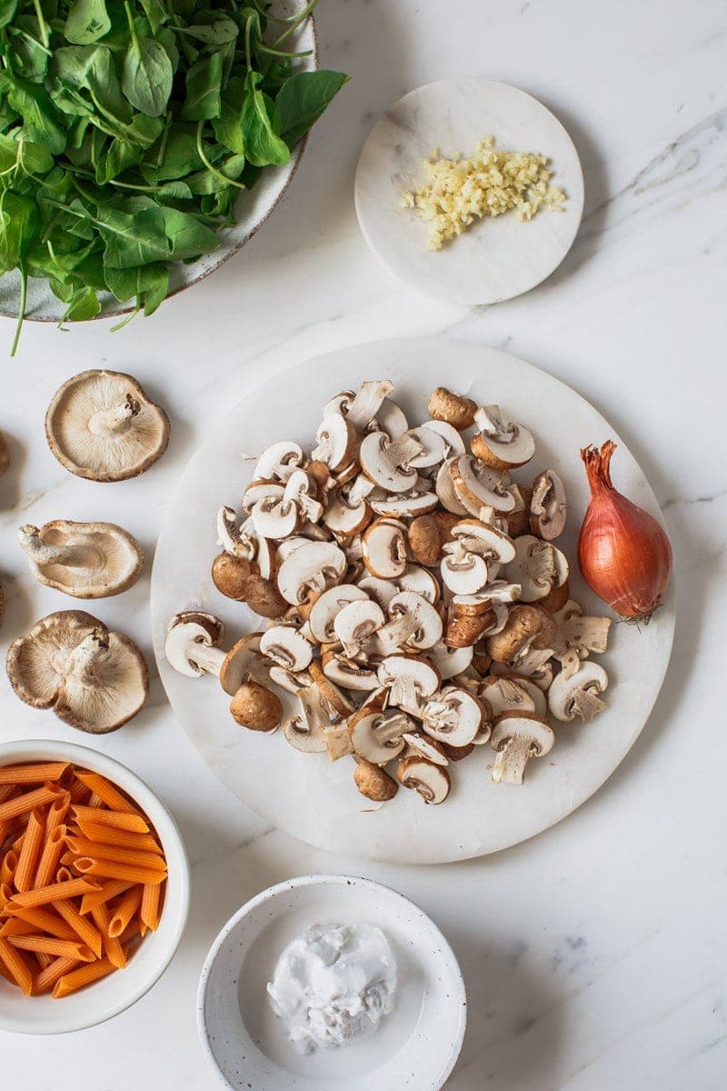 Fresh ingredients for creamy mushroom pasta laid out on the bench - mushrooms, arugula, shallot, garlic, penne, coconut cream