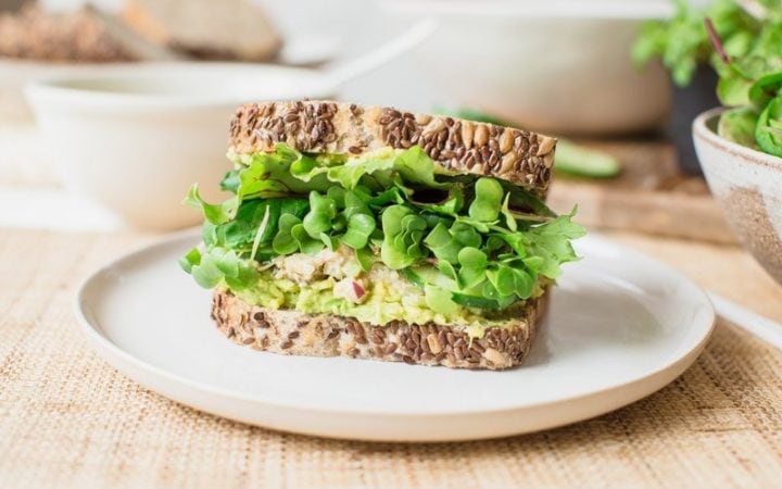 A sandwich made from rustic bread jam packed with fresh greens, avocado and a smashed chickpea filling. On a plate ready to eat.