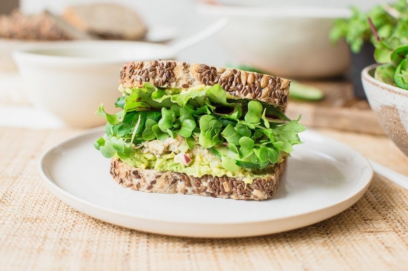 A sandwich made from rustic bread jam packed with fresh greens, avocado and a smashed chickpea filling. On a plate ready to eat.