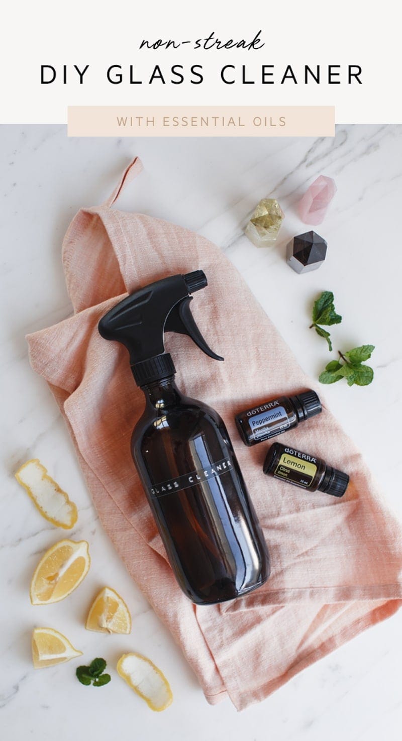 Hero shot of the glass cleaner on a linen napkin, surrounded by lemon peel, fresh mint, and a few bottles of essential oils, the recipe title is written over the top of the image