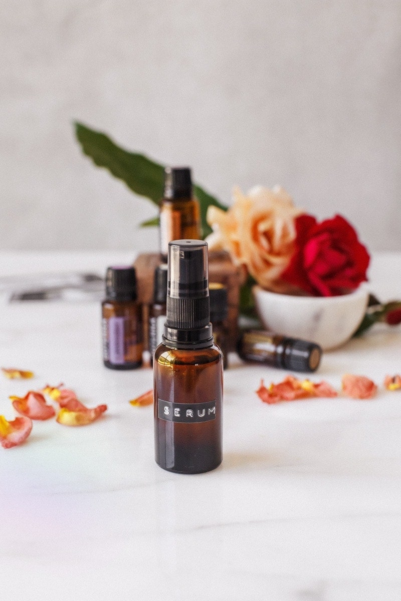 Glass serum pump bottle in the foreground with essential oils and fresh decorative roses in soft focus in the background