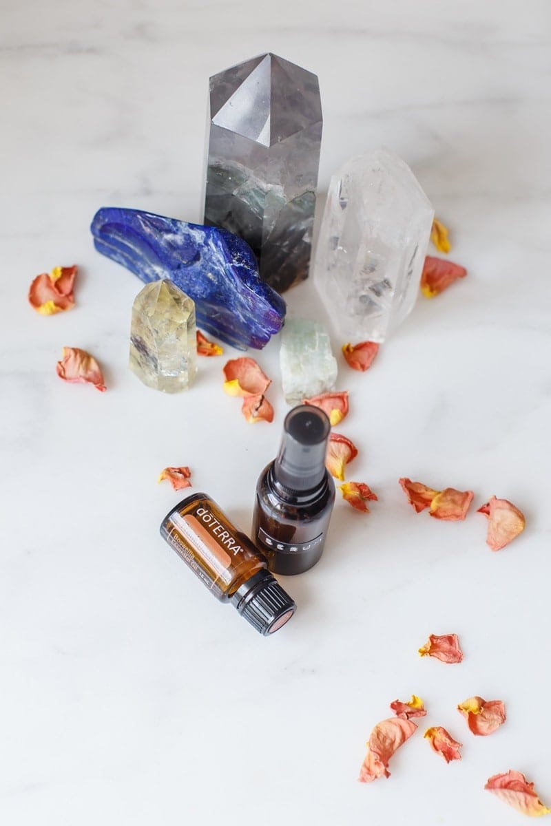 Overhead shot of the serum and a bottle of frankincense on a marble surface, with decorate crystals and rose petals beside them