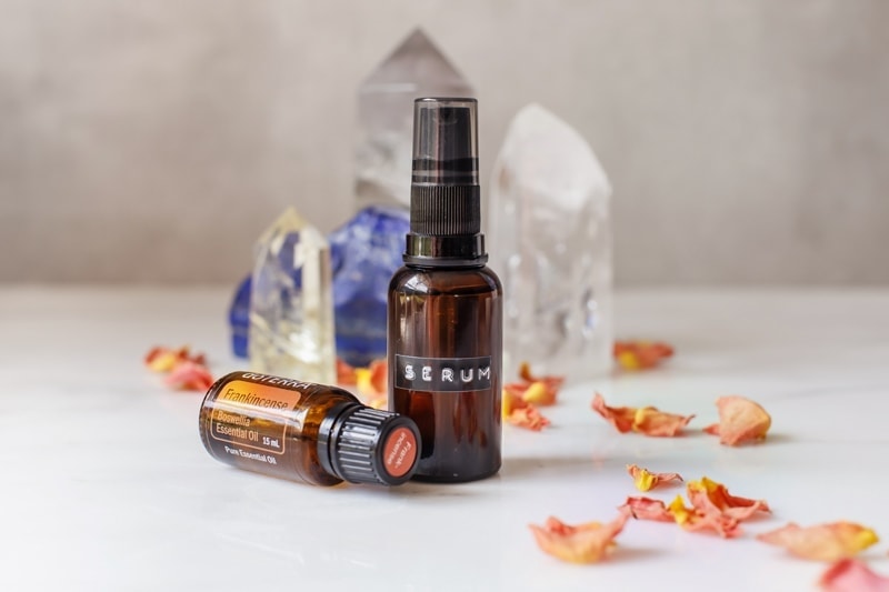 A small amber glass serum pump beside a bottle of Frankincense, with dried rose petals and a crystal in the background