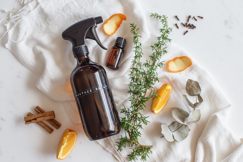 Amber glass spray bottle on a marble counter, surrounded by rosemary sprigs, orange peel, cinnamon sticks and clove