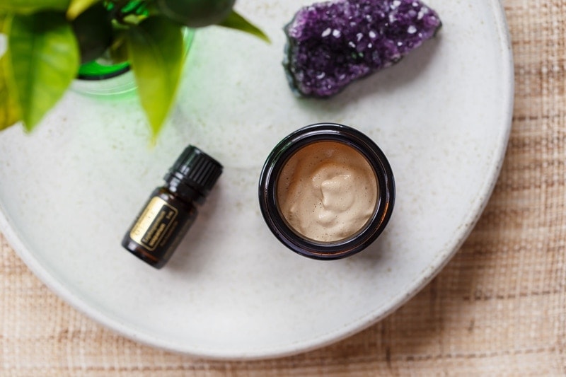 Close up of a pottle of homemade face cream on a ceramic plate with a small bottle of Manuka essential oil beside it