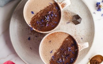 Two mugs filled with a 'shroom hoot chocolate, dusted with cacao and topped with dried blue cornflower petals