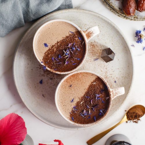 Two mugs filled with a 'shroom hoot chocolate, dusted with cacao and topped with dried blue cornflower petals