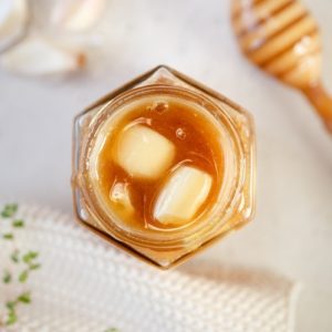 Small jar of Manuka honey with cloves of peeled and bruised garlic throughout - honey dipper in the background.