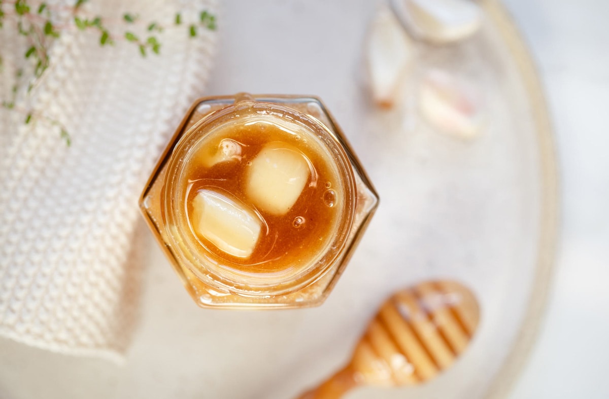Over head shot of a hexagonal jar of honey with fermented garlic cloves inside. Bubbles are at the surface.