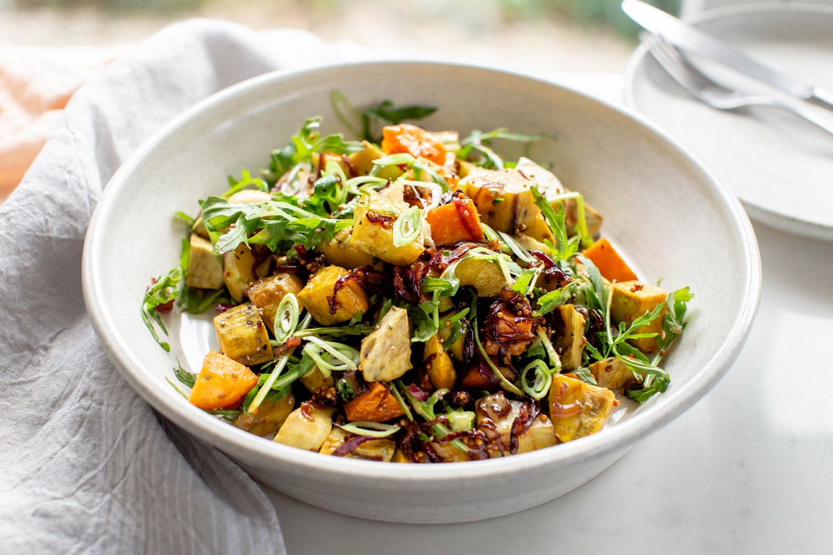 Roast kumara salad in a ceramic serving bowl topped with caramelised onions. Linen napkin rests underneath it.