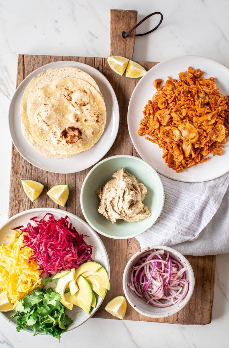 A wooden board full of ingredients used to make vegan tacos with - hummus, fresh beets, avocado, pickled onions, tempeh meat, tortillas!