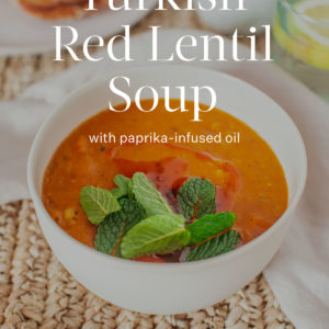 Beautifully styled bowl of red lentil soup on a woven placemat topped with mint and paprika oil, text over the top reads 'Turkish Red Lentil Soup.'