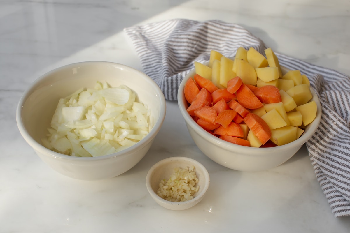 Prepped vegetables - a bowl of diced onion, a other of diced potato and carrot, and a third of minced garlic.