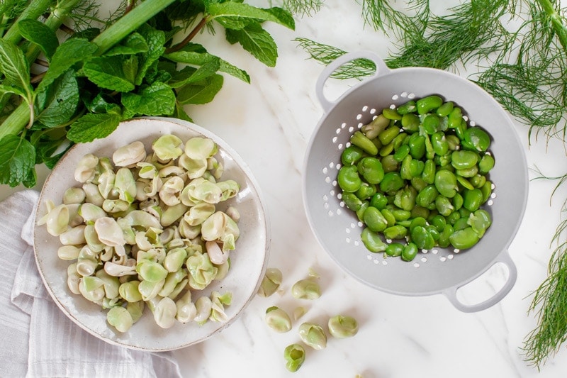 Broad beans that have been popped out of their pods, in a colander, with the skins in a bowl beside them