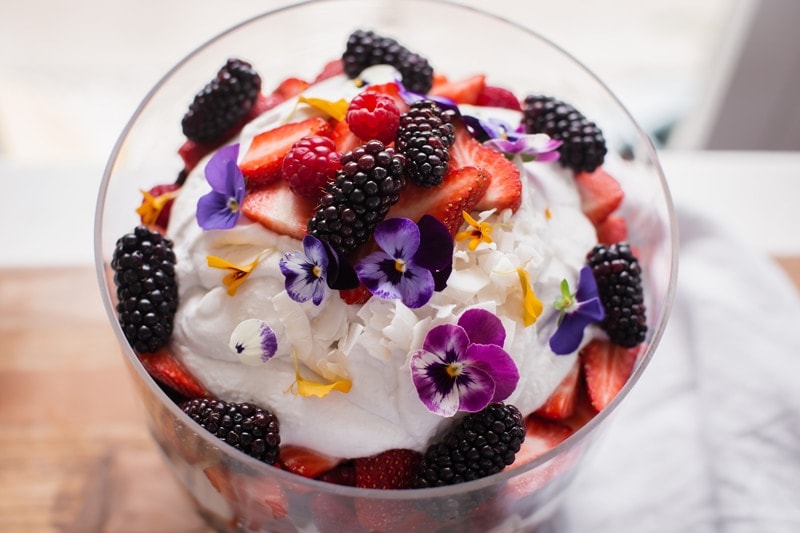 Close up of the top of a trifle with a mound of whipped coconut cream, raspberries, strawberries, blackberries and flowers