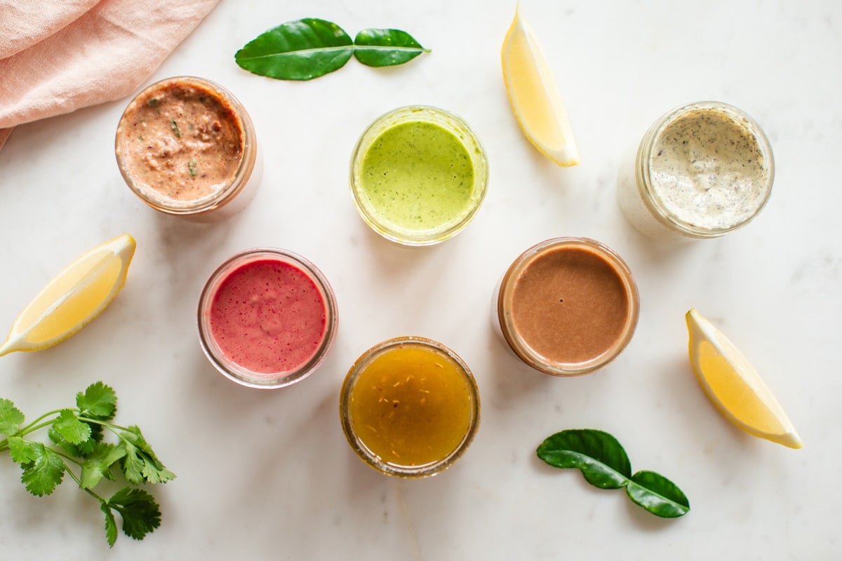 Overhead shot of 6 healthy salad dressing recipes in open jars, showing off the varied vibrant colours