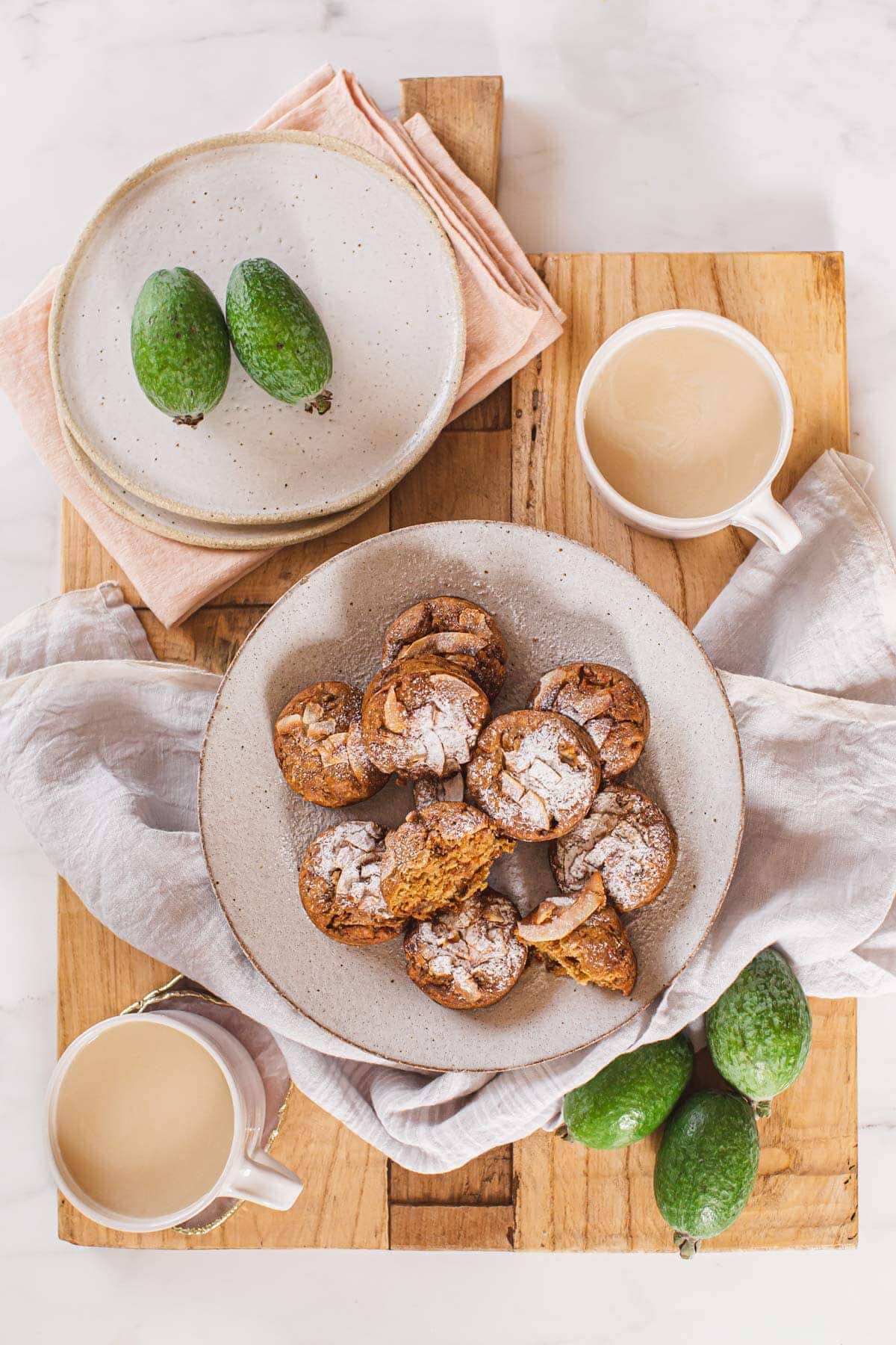 Overhead shot of muffins on a plate, on a wooden board, with fresh feijoas and two cups of milky chai ready to serve