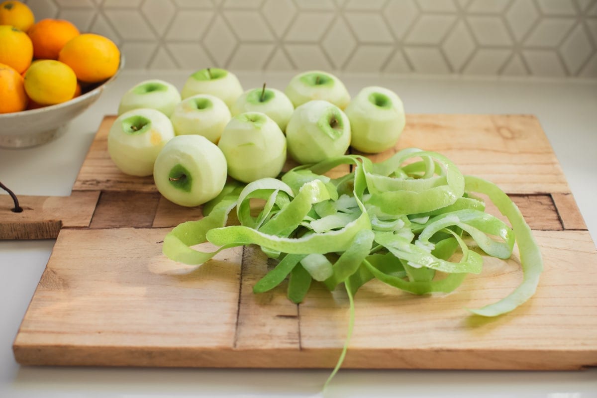 10 peeled Granny Smith apples on a wooden board, with a pile of the apple peel to the side.