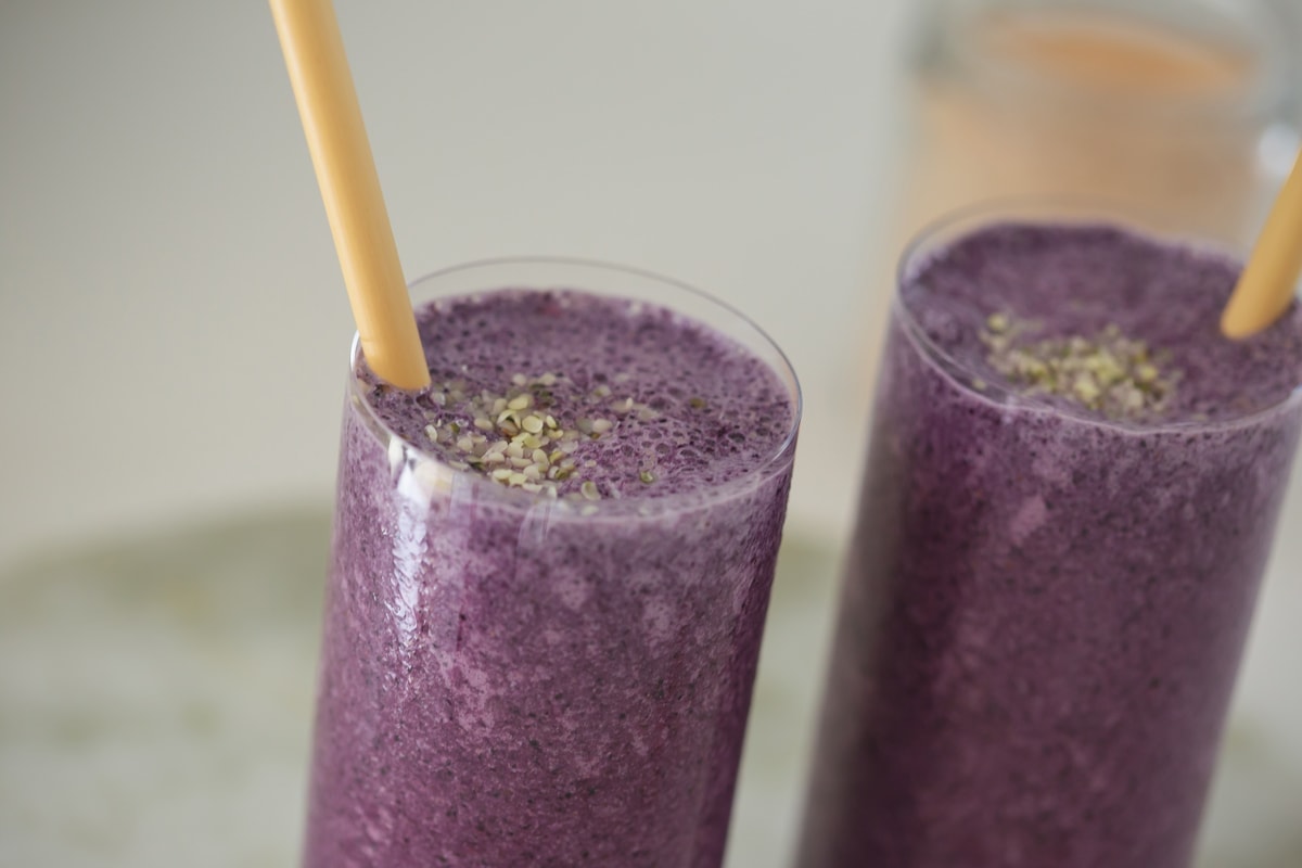 Close up of the tops of two tall bright blue-purple coloured smoothies with wooden straws and hemp seeds sprinkled over the top.